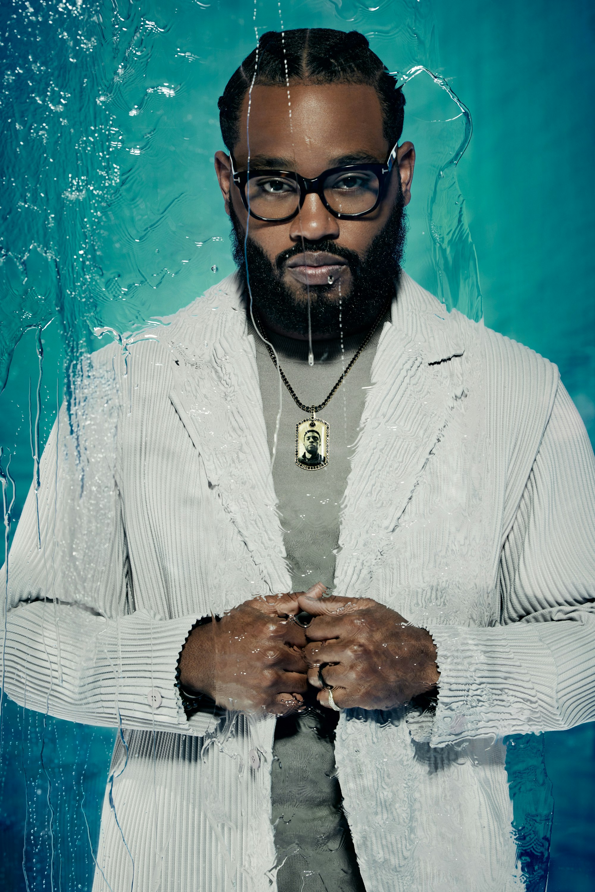 Portrait of a bearded man with glasses wearing a striped blazer and a necklace, posing in front of a textured blue background.