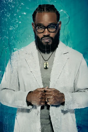 Portrait of a bearded man with glasses wearing a striped blazer and a necklace, posing in front of a textured blue background.