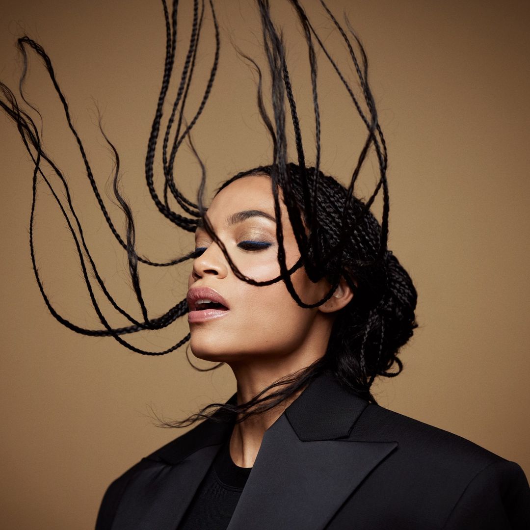 Close-up portrait of a woman with braided hair tossed in motion against a warm neutral background wearing a black blazer