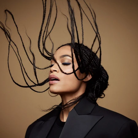 Close-up portrait of a woman with braided hair tossed in motion against a warm neutral background wearing a black blazer