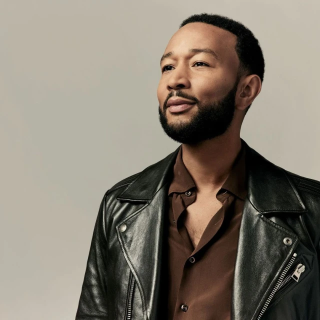 Portrait of John Legend wearing a black leather jacket and brown shirt against a grey background, looking off to the left.