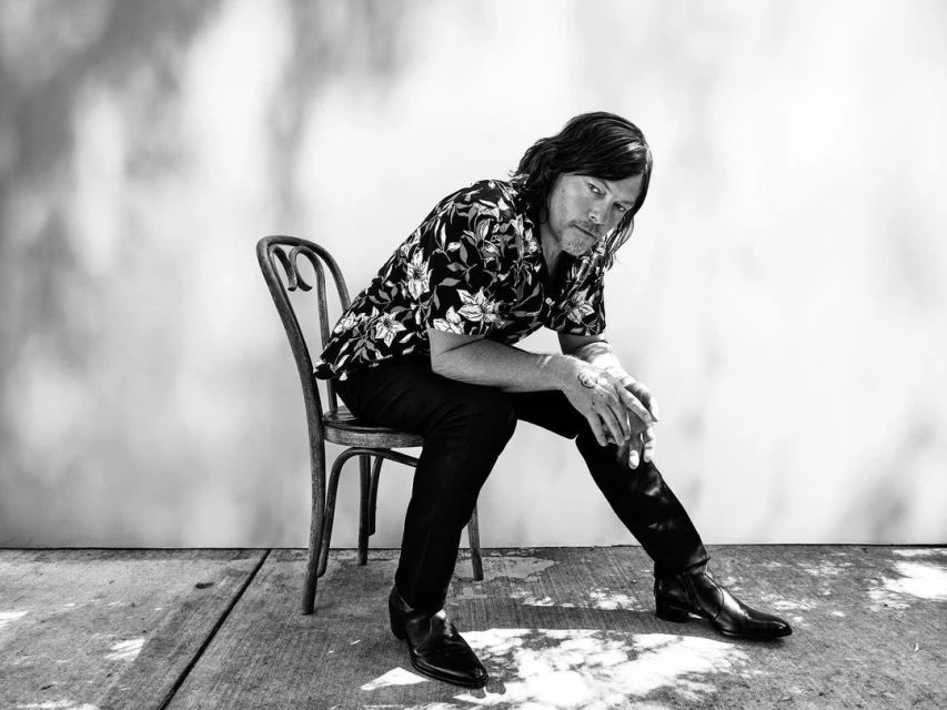 Monochrome portrait of a man with long hair wearing a floral shirt, sitting on a chair outdoors in dappled sunlight.