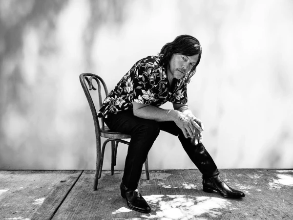 Monochrome portrait of a man with long hair wearing a floral shirt, sitting on a chair outdoors in dappled sunlight.