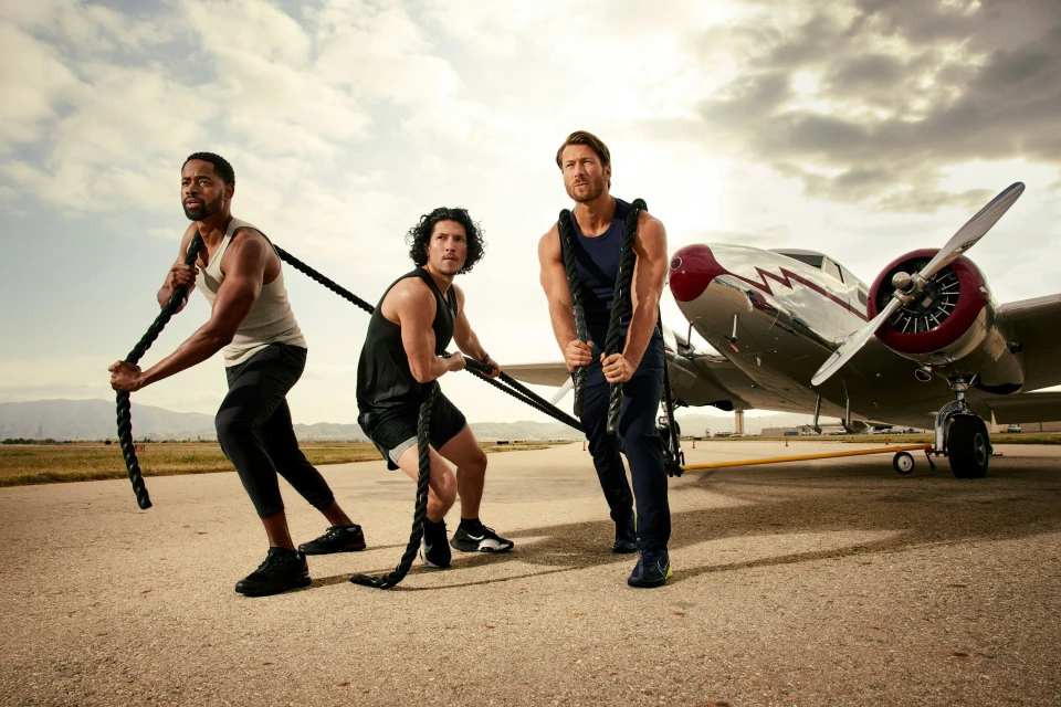 Three adult men pull thick ropes in unison on an airport runway with a vintage airplane in the background under a cloudy sky.