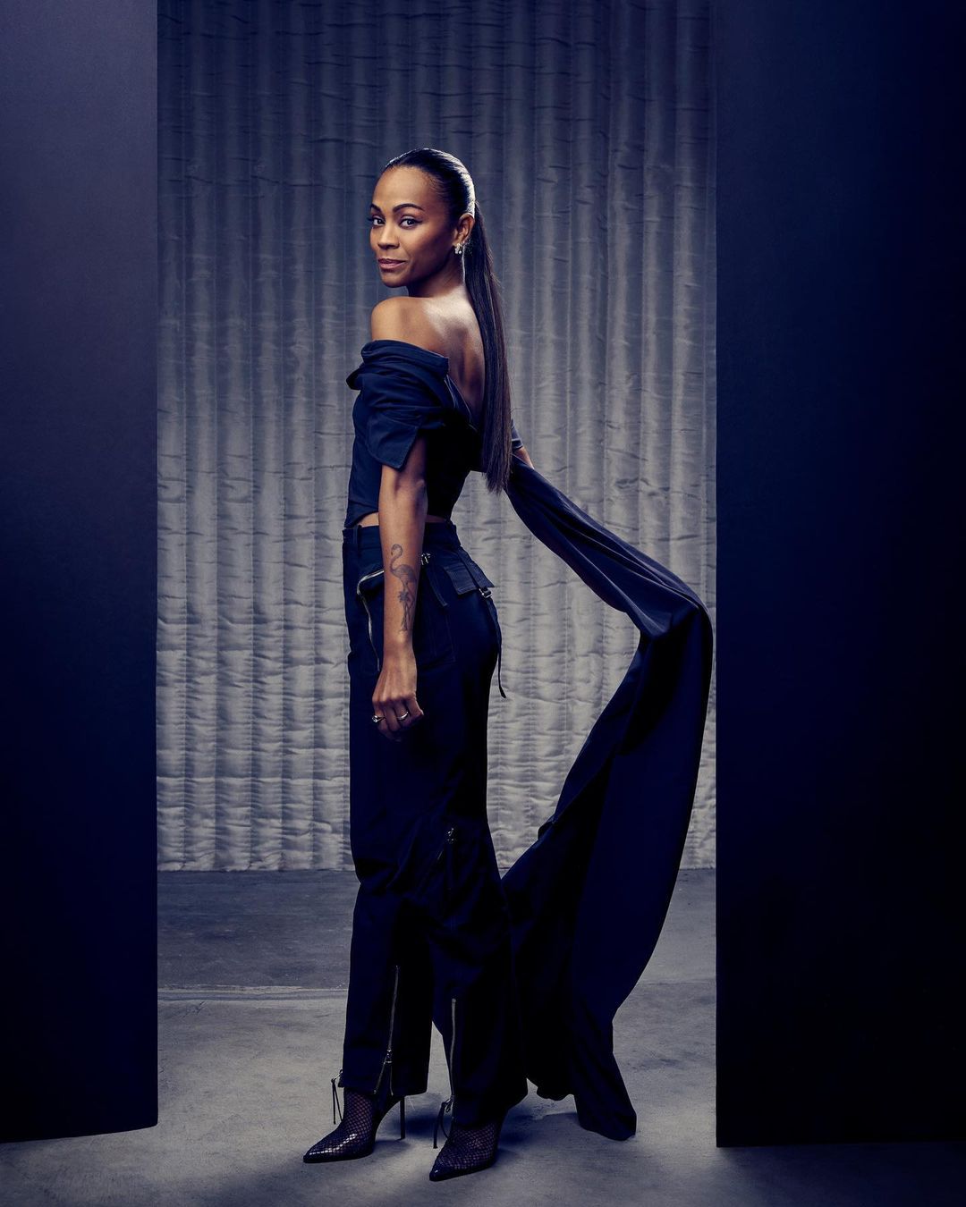 Zoe Saldana poses in off-the-shoulder black top and pants with a long train, side view in a photo studio with a textured backdrop.
