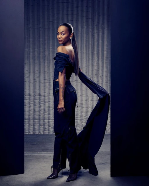 Zoe Saldana poses in off-the-shoulder black top and pants with a long train, side view in a photo studio with a textured backdrop.