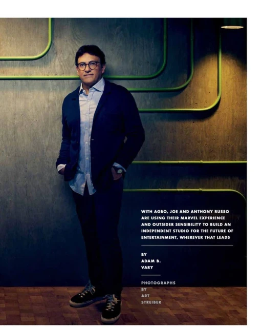 Portrait of Anthony Russo in front of a patterned wall, wearing a dark blue blazer, light blue shirt, dark pants, and casual shoes.