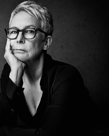 Black and white portrait of mature adult female, Jamie Lee Curtis, with glasses, wearing a dark open-collar shirt, hand to face.