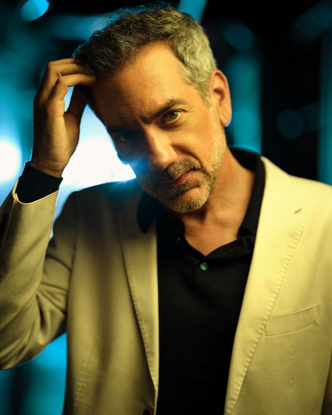 Portrait of a man with graying hair, wearing a blazer and black shirt, thoughtfully touching his head in soft, teal-toned lighting.