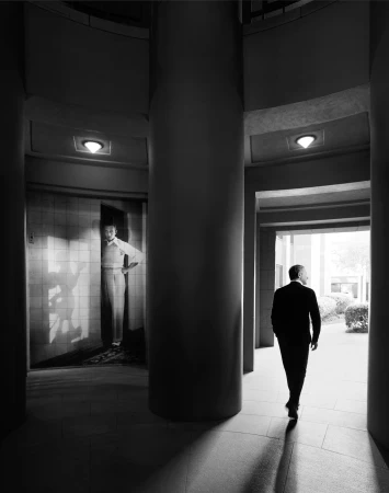 Monochrome image featuring a man in a suit walking through a colonnade, with another man visible in an opening on the left.
