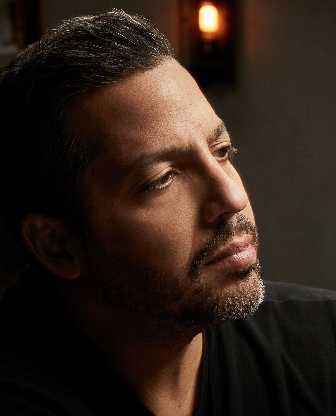Headshot portrait of an adult man with short hair and a graying beard looking off-camera, illuminated by soft studio lighting.