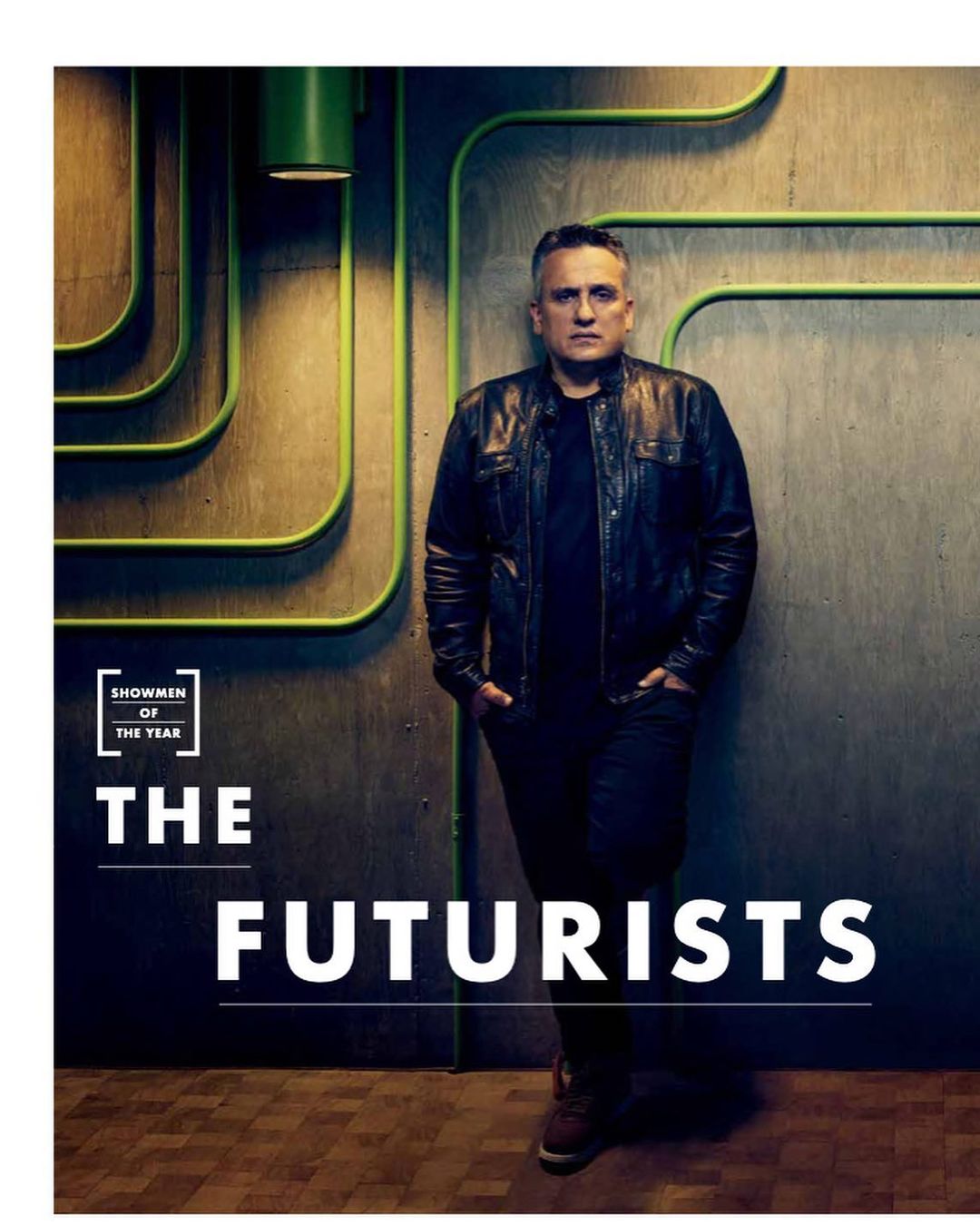 Portrait of a man in a black jacket standing against a textured wall with graphic neon tubing and magazine cover graphics.