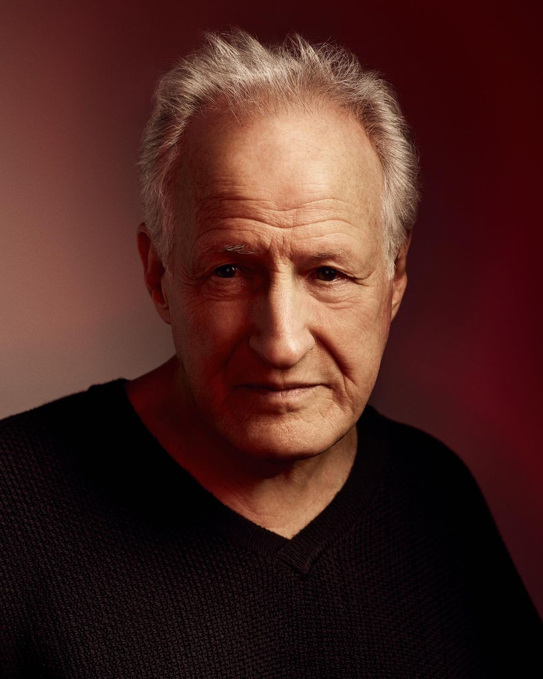 Portrait of an older adult male with short grey hair wearing a dark V-neck shirt, set against a blurred red background in eye-level medium shot.