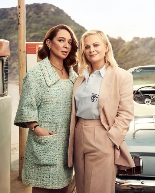 Two adult women, Amy Poehler and Maya Rudolph, pose near a vintage car and gas station against a hilly background wearing fashionable attire.