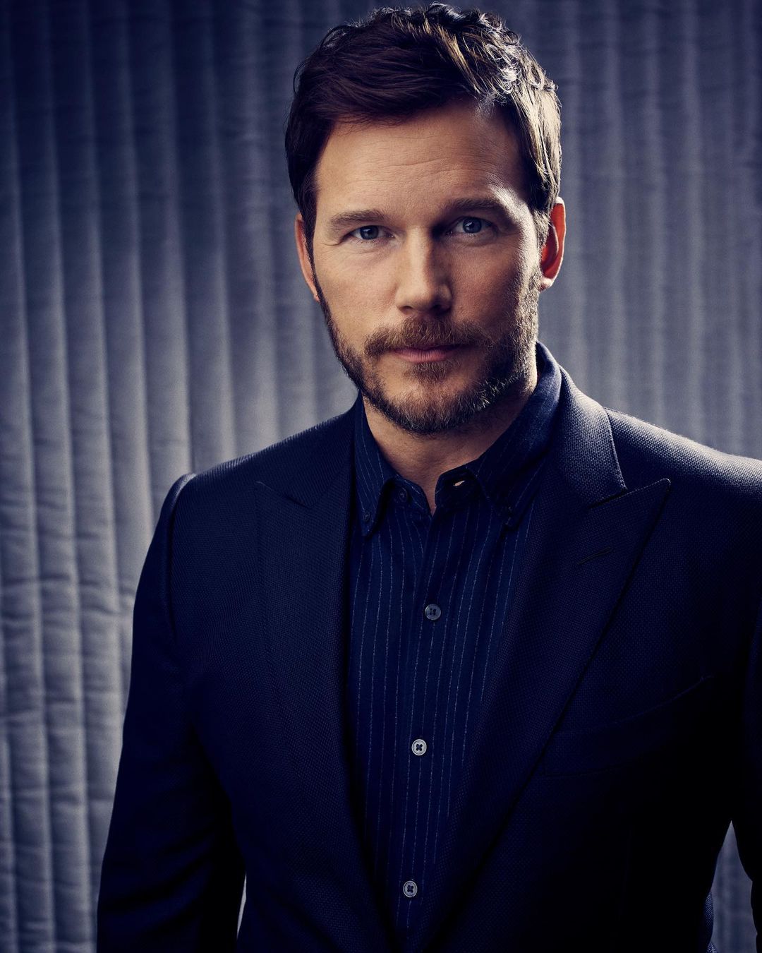 Portrait of Chris Pratt with a neatly groomed beard wearing a blue shirt and a dark suit jacket against a grey patterned backdrop.