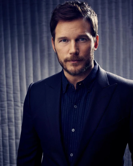 Portrait of Chris Pratt with a neatly groomed beard wearing a blue shirt and a dark suit jacket against a grey patterned backdrop.