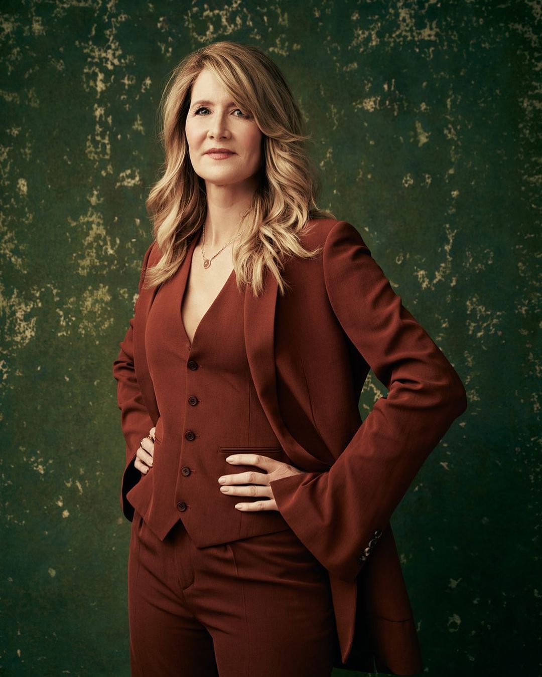 A medium portrait of an adult female with long blonde hair wearing a red-brown pant suit with her hands on her hips against a green mottled background.