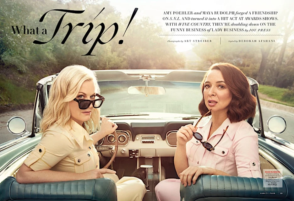 Amy Poehler and Maya Rudolph pose in a vintage convertible car, with a road and trees blurred in the background, in a Vanity Fair photograph.