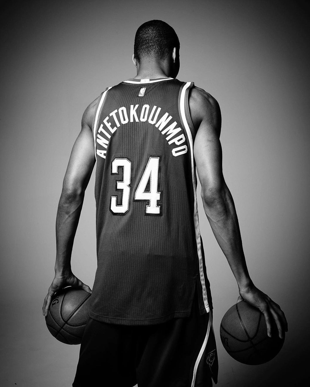 Black and white studio shot of a muscular athlete in sports jersey and shorts holding two basketballs, back to camera.