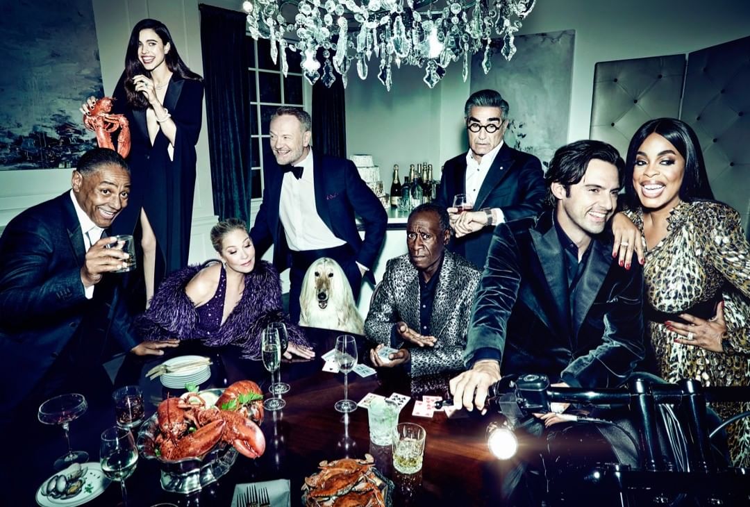 Group of celebrities in formal wear around a table laden with seafood, with a dog and motorcycle, posing for a portrait in a luxurious setting.