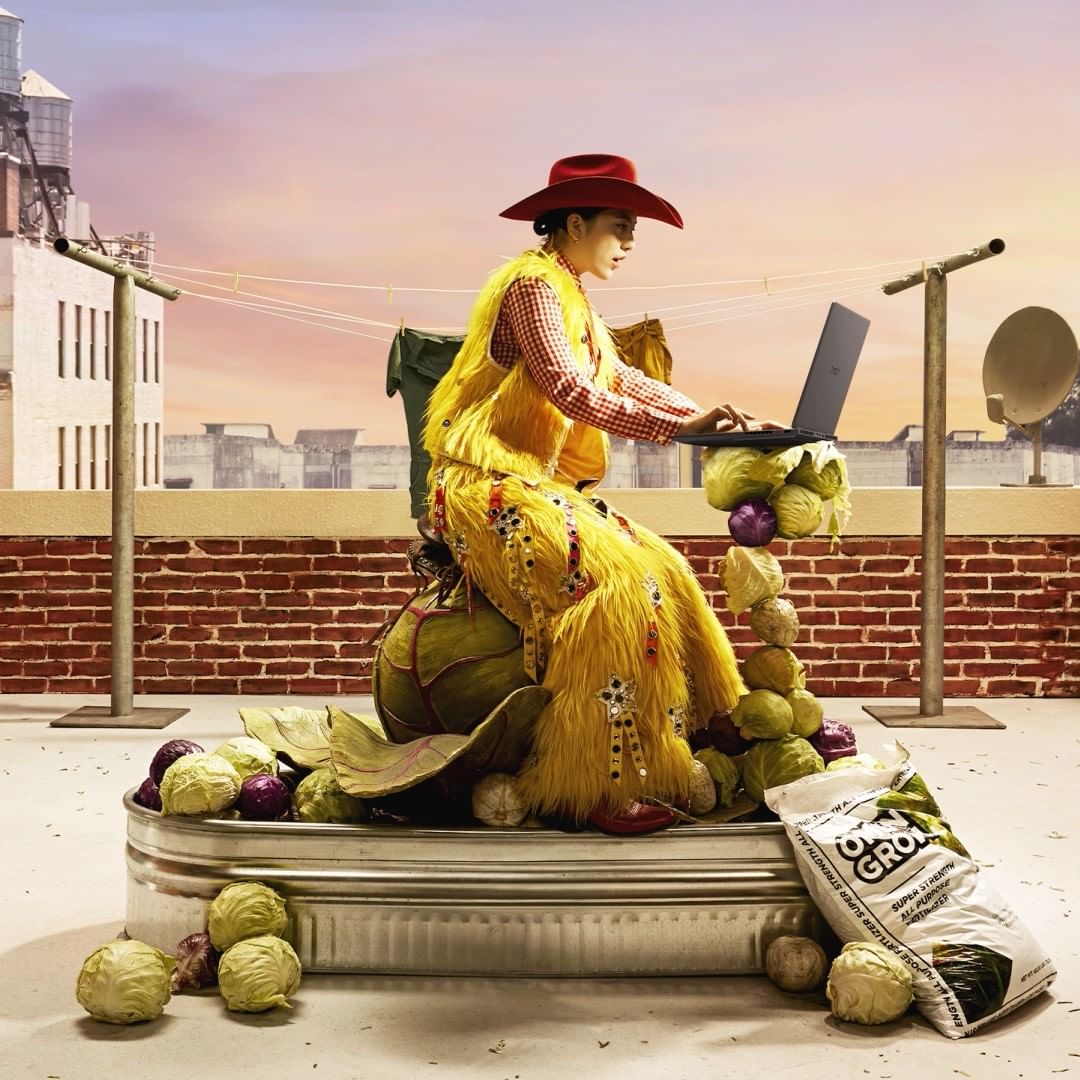 Woman in vibrant yellow outfit and red hat sits atop a large green cabbage on a rooftop, using a laptop surrounded by produce.