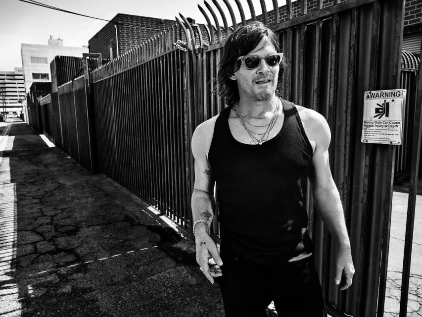 Black and white portrait of an adult male in sunglasses and a tank top, standing outdoors near a metal fence in an urban setting.