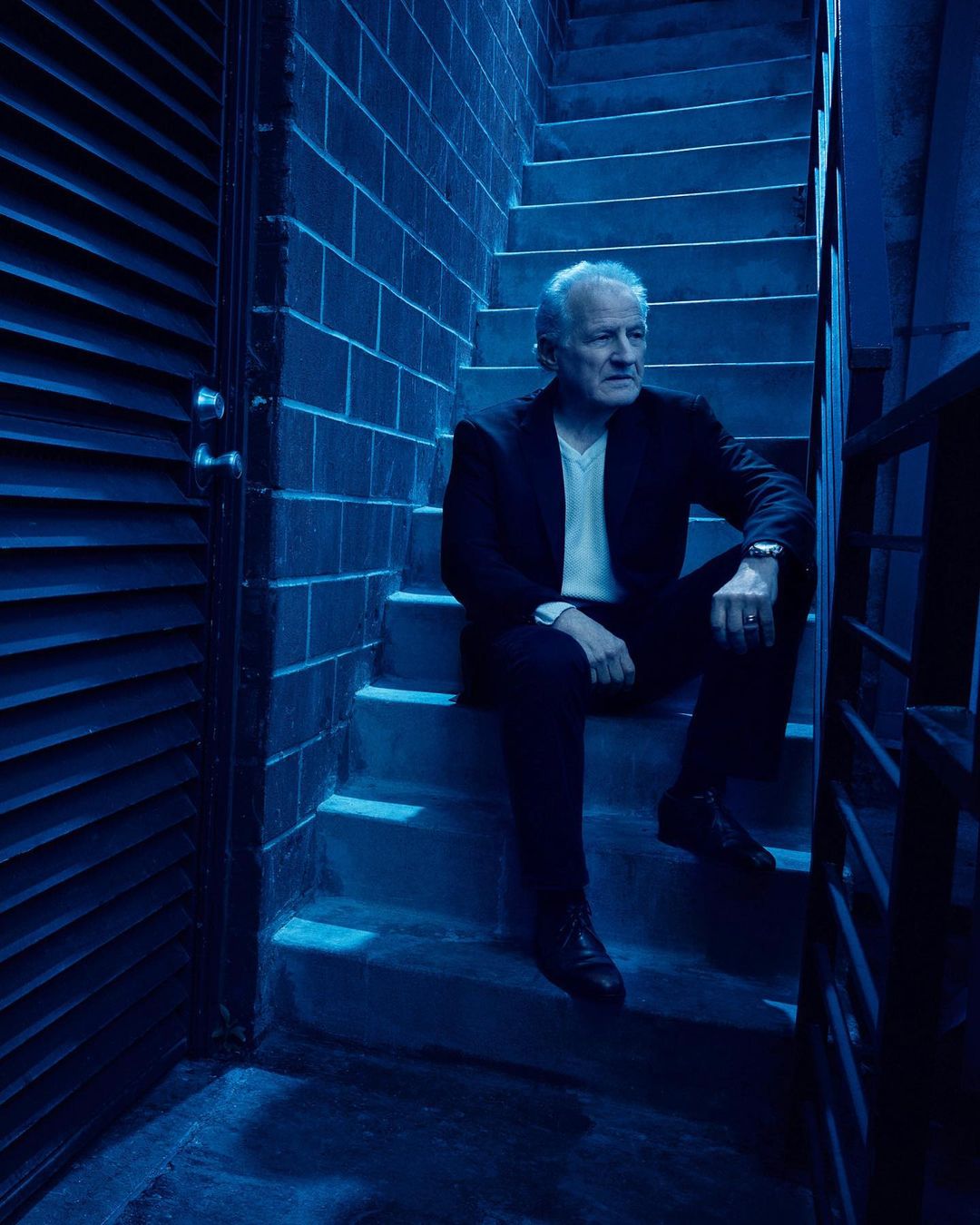 An older adult male sits on blue concrete stairs in a black suit jacket and dark pants looking off-camera, moody lighting.