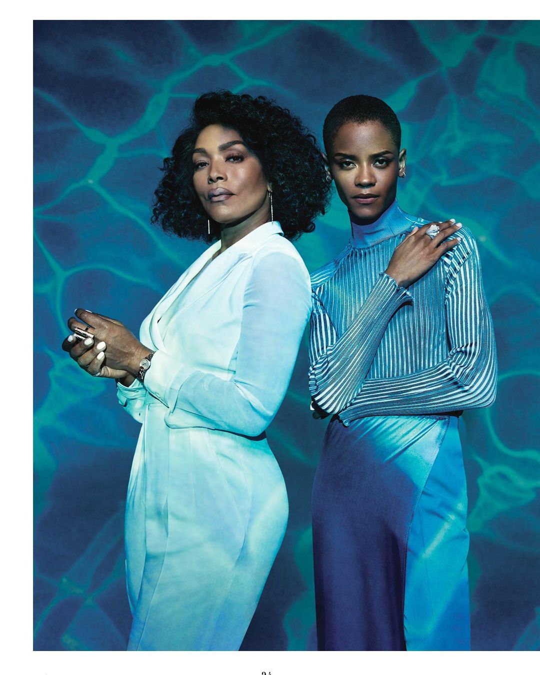 Two women stand in a studio portrait with a textured blue background. One woman wears a white suit, and the other wears a striped top and skirt.