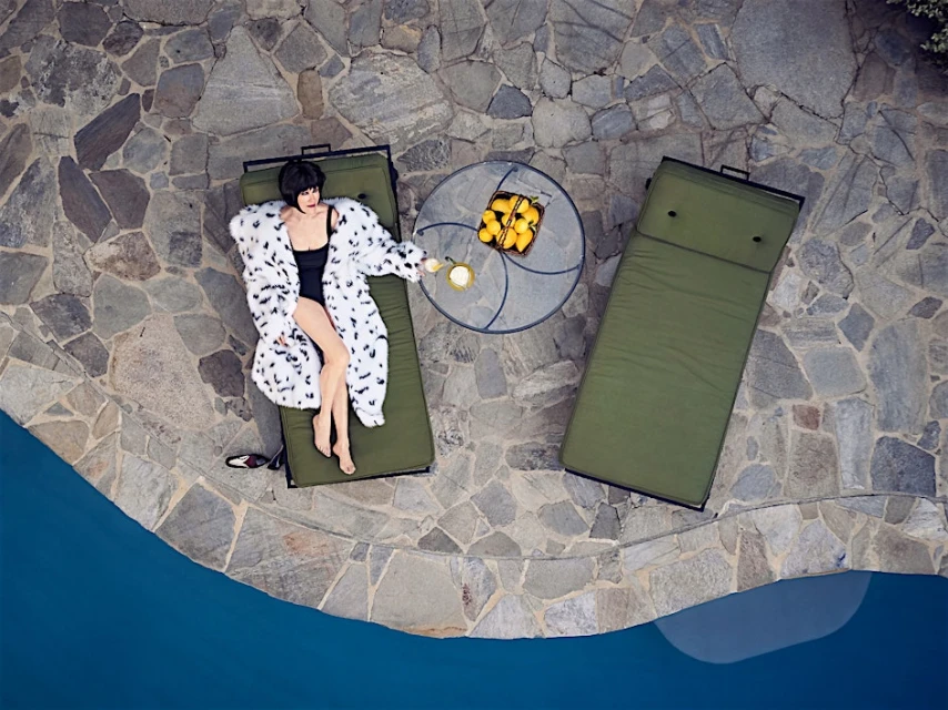 Overhead view of a woman in a spotted coat relaxing on a lounge chair by a pool, with lemons and an empty chair nearby.