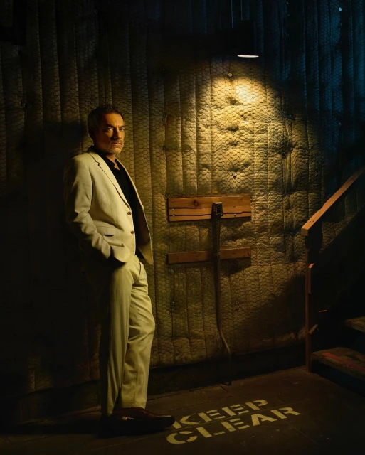 Portrait of a mature man in a light-colored suit standing against a textured wall in subdued lighting with 'Keep Clear' stenciled on the floor.
