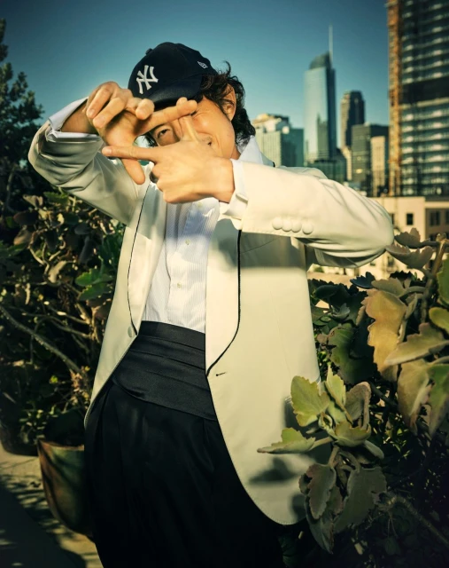 Man in a white suit jacket and baseball cap stands on a rooftop with city buildings in the background, framing his face with his hands.