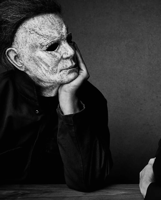 Person in Michael Myers mask with a sullen expression, resting head on hand at a table with a dark background.