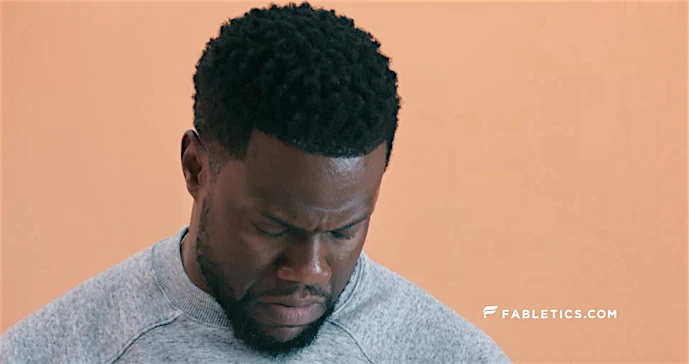 Close-up portrait of Kevin Hart wearing a grey sweatshirt against a solid peach background.