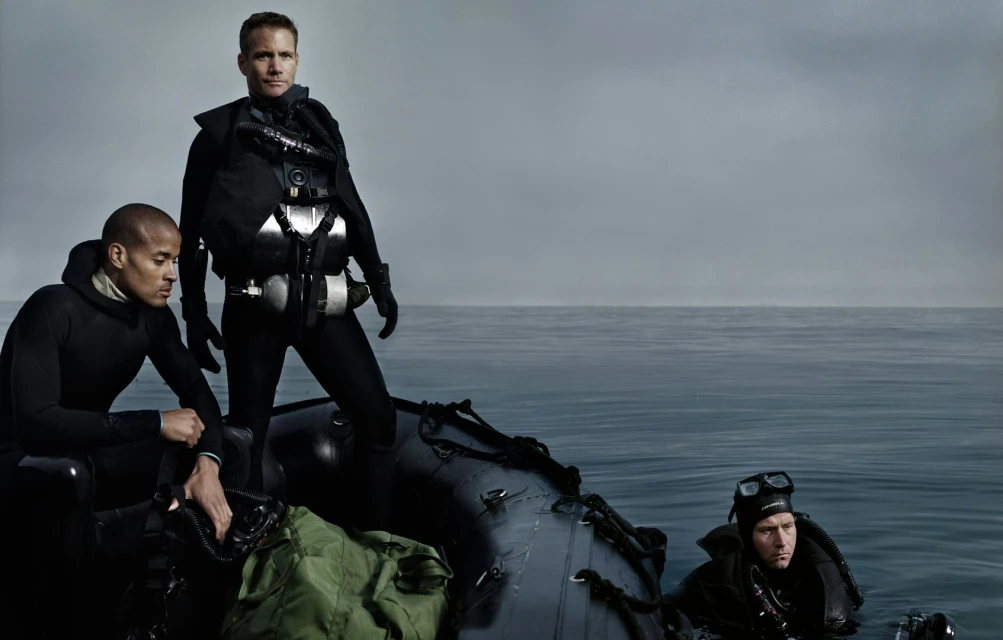 Three divers in black wetsuits on an inflatable boat in the ocean under a cloudy sky, preparing for a dive, focused and ready.