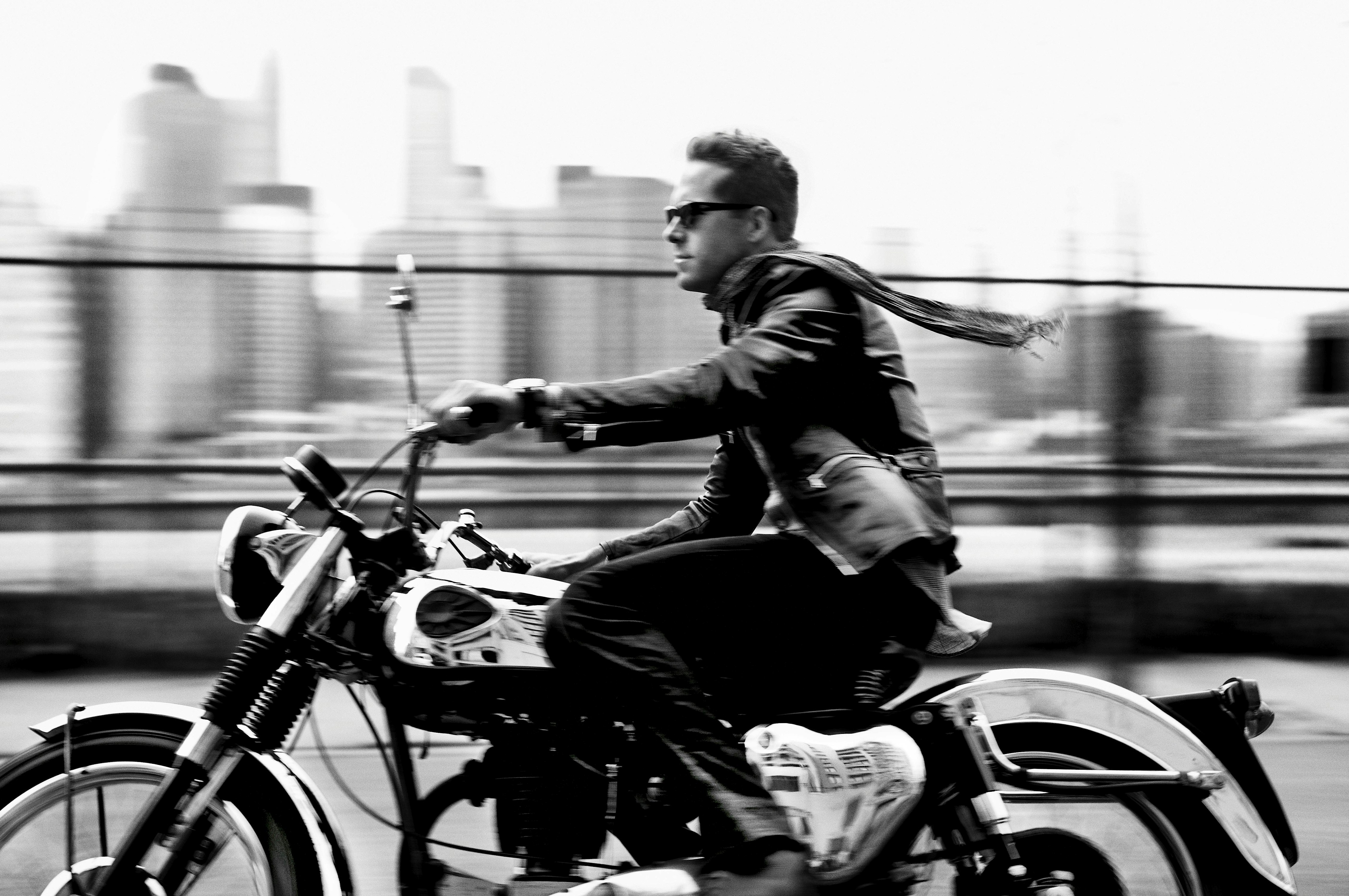 Monochrome shot of a man riding a motorcycle with a city skyline in the background, conveying motion and urban style.
