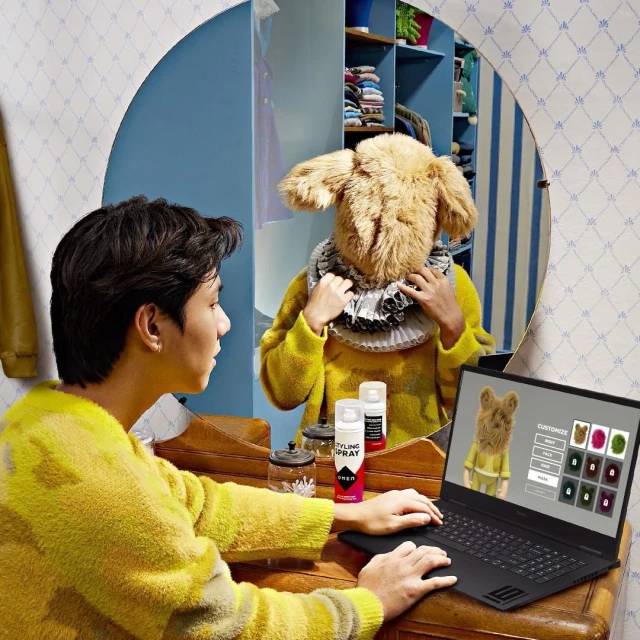 Person sitting at a desk wearing a yellow sweater, using a laptop, with a mirror reflection of a person wearing a costume.