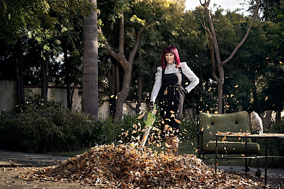 Woman with pink hair uses a leaf blower on a pile of leaves in an outdoor setting, with trees, chair, and owl statue.