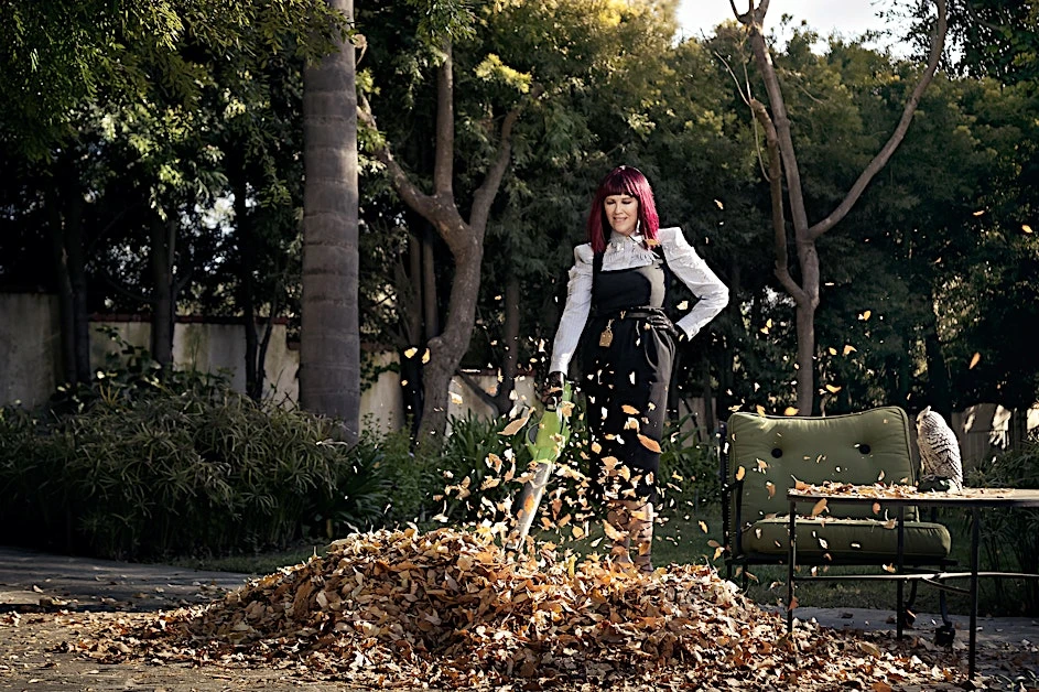 Woman with pink hair uses a leaf blower on a pile of leaves in an outdoor setting, with trees, chair, and owl statue.