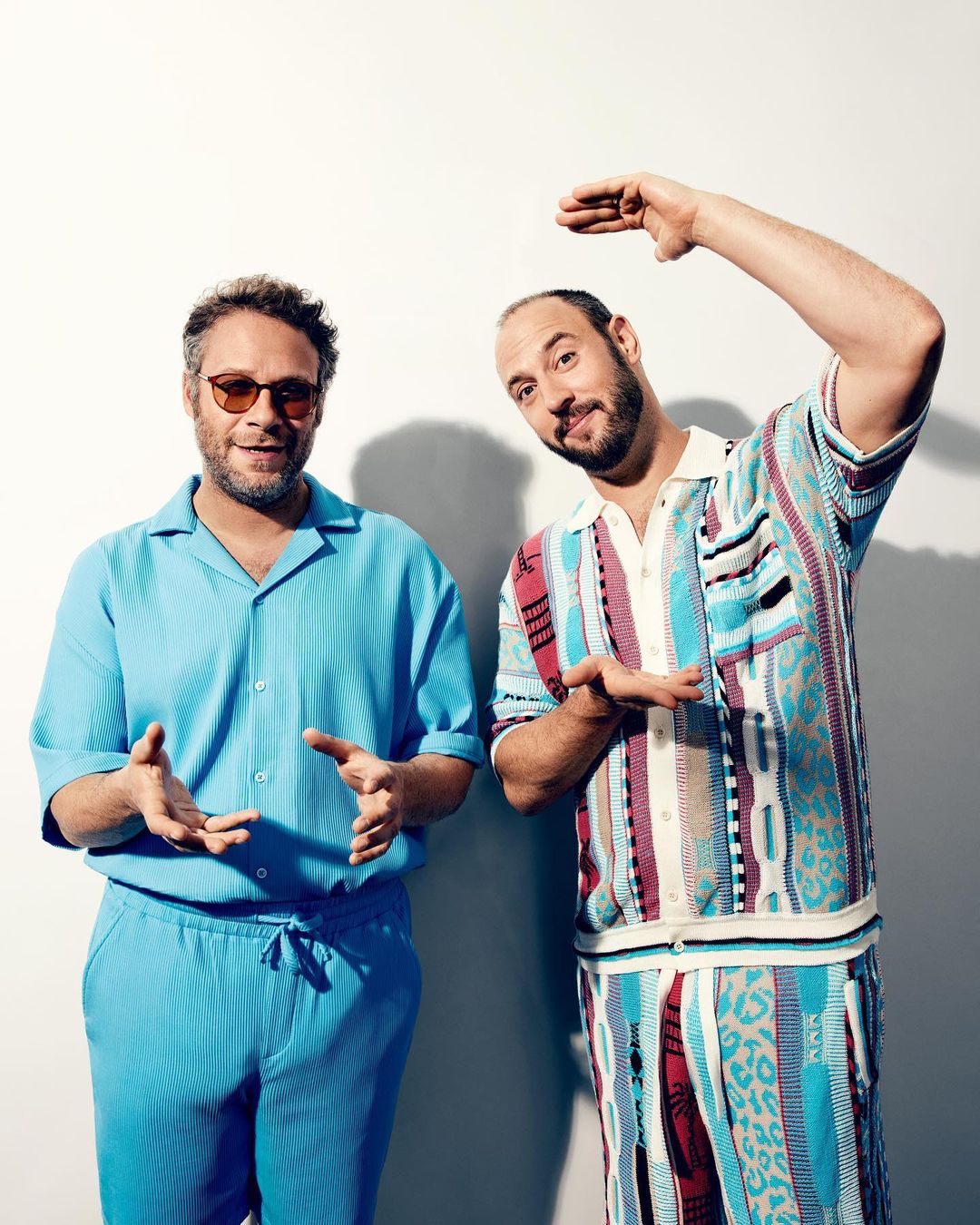 Two adult males pose indoors against a white wall, wearing casual outfits in a medium shot, displaying contemporary fashion.