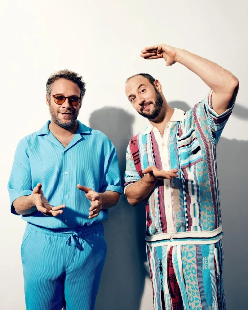 Two adult males pose indoors against a white wall, wearing casual outfits in a medium shot, displaying contemporary fashion.