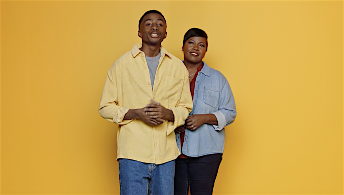 Two people, an adult man and woman, stand close together smiling in front of a yellow backdrop in a medium shot.