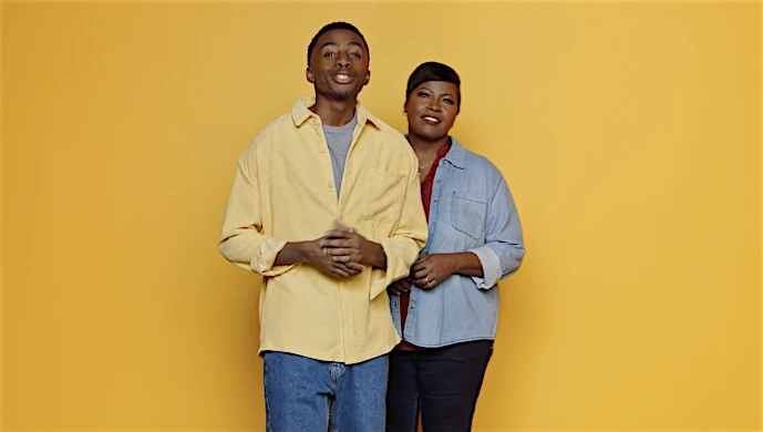 Two people, an adult man and woman, stand close together smiling in front of a yellow backdrop in a medium shot. 