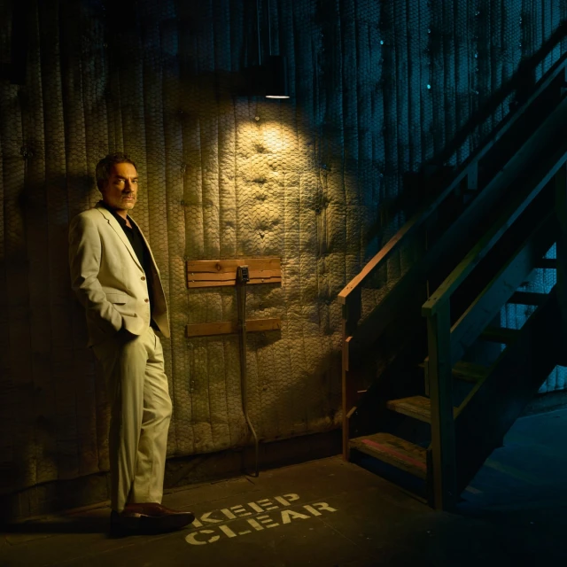 Man in a cream suit stands indoors next to a wall with weathered texture and a staircase, lit by a spotlight. The lighting is dramatic.