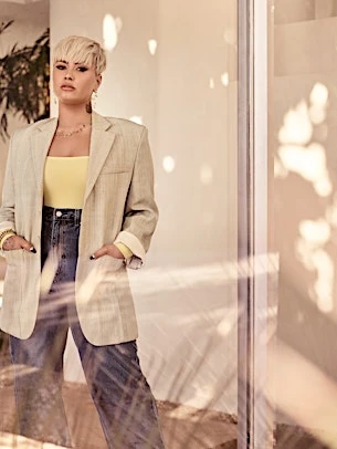 Adult woman in oversized tan blazer, yellow top, and jeans stands in a sunlit interior with her hands in her blazer pockets.