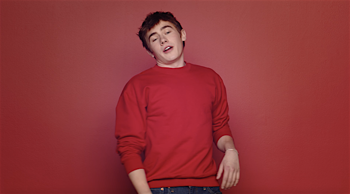 Portrait of a young man in a red sweater standing against a matching red background. He is looking at the camera and talking.