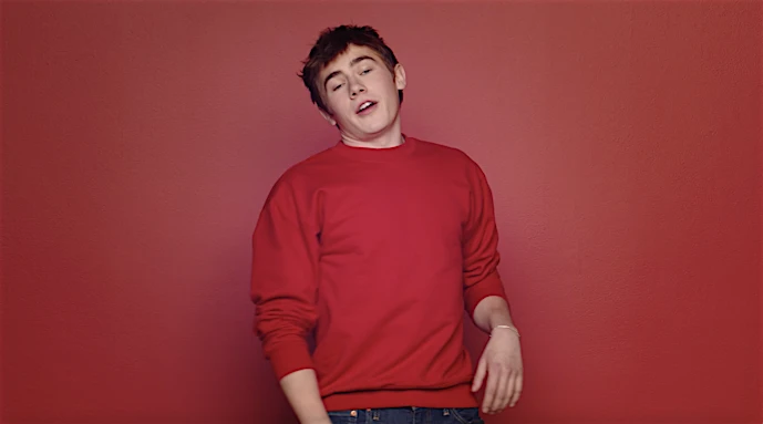 Portrait of a young man in a red sweater standing against a matching red background. He is looking at the camera and talking.