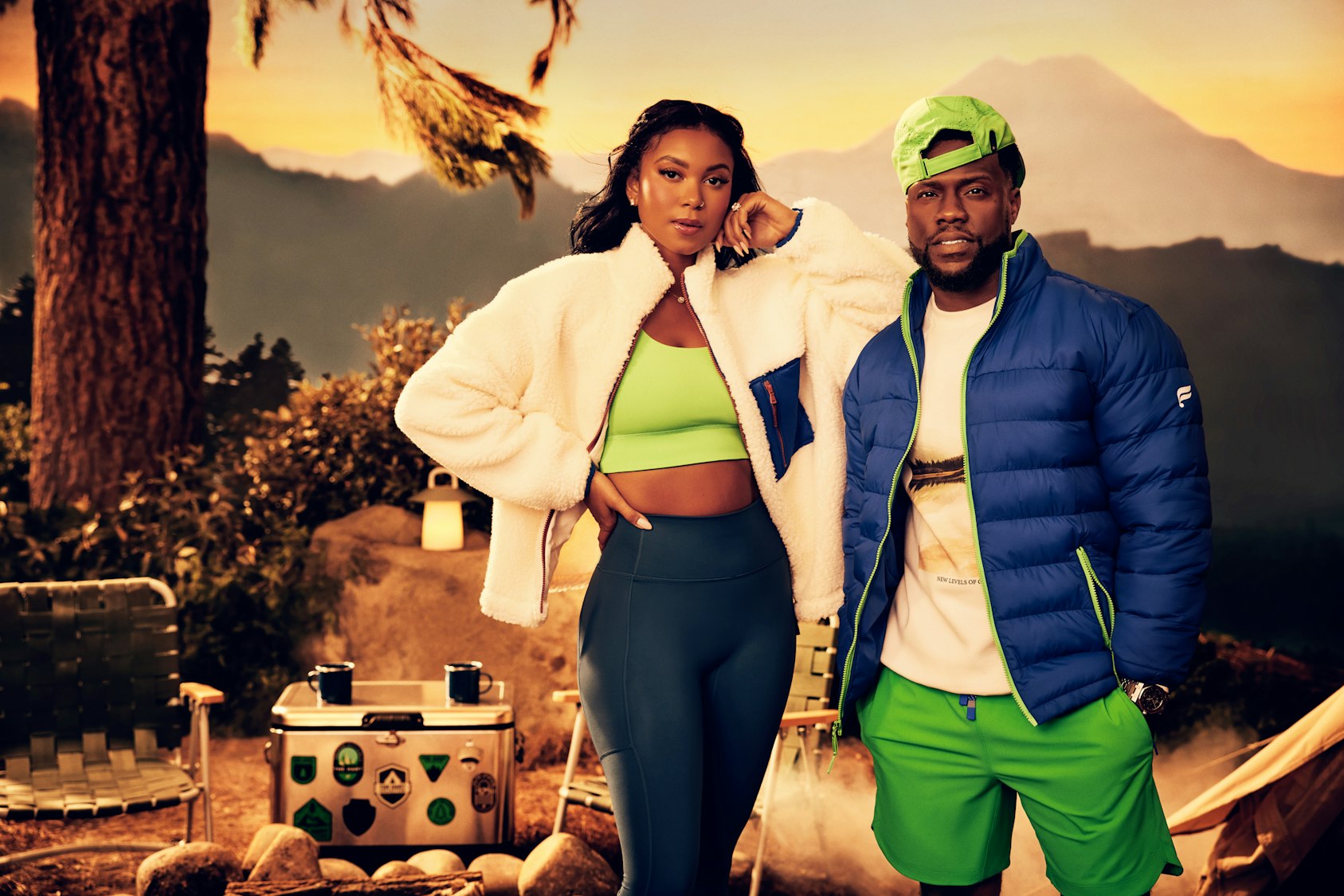 Two athletic adults, one male and one female, pose in a staged outdoor scene with camping props and painted mountain backdrop.