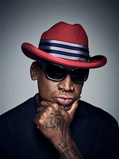 Portrait of an adult male wearing a red hat and sunglasses, hand on chin, tattoos visible on hand, set against a grey background.