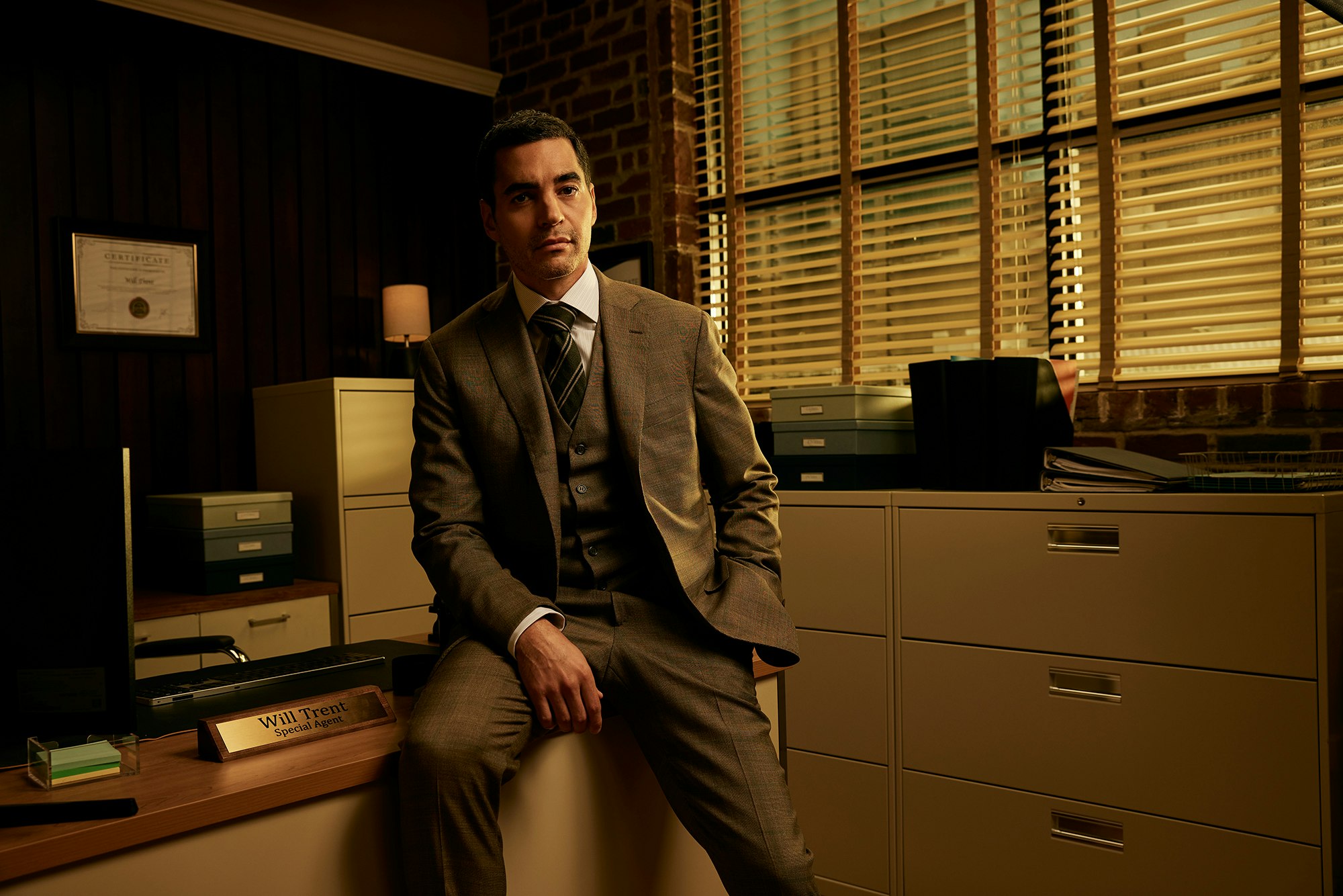 Man in suit sits on desk in office with filing cabinets. Natural lighting, certificate on wall. Calm facial expression.  