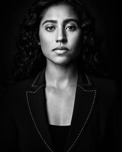 Portrait of an adult woman with curly hair wearing a black blazer with white stitching details, set against a dark background.